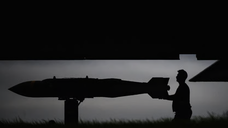 Silhouette of a military personnel loading a large missile or bomb under an aircraft wing at dusk.