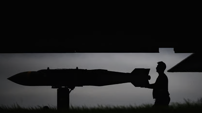 Silhouette of a military personnel loading a large missile or bomb under an aircraft wing at dusk.