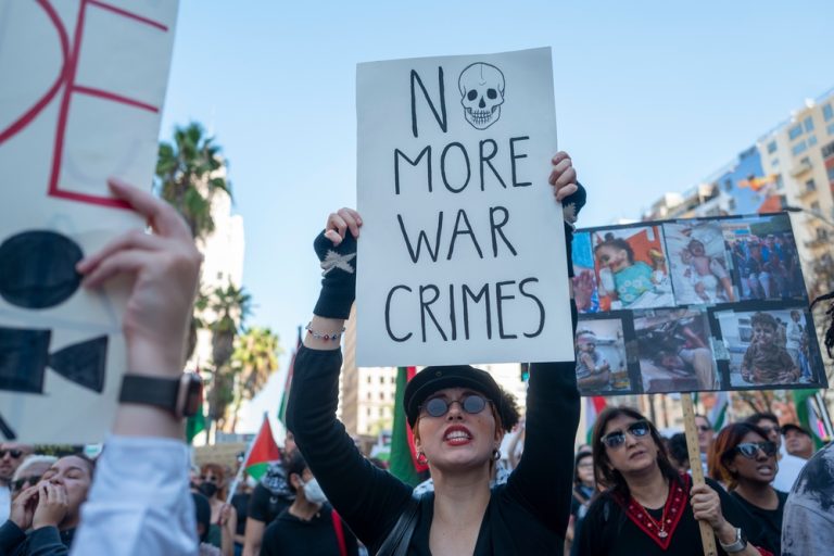 Protester holding a sign that says "NO MORE WAR CRIMES" with a skull illustration during a street demonstration.