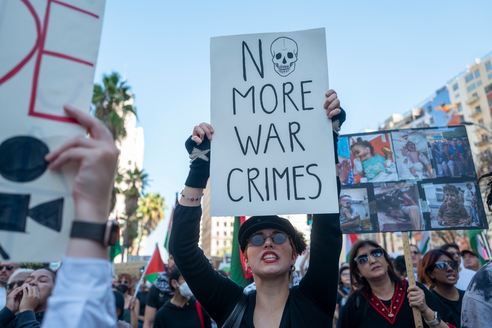 Protester holding a sign that says "NO MORE WAR CRIMES" with a skull illustration during a street demonstration.