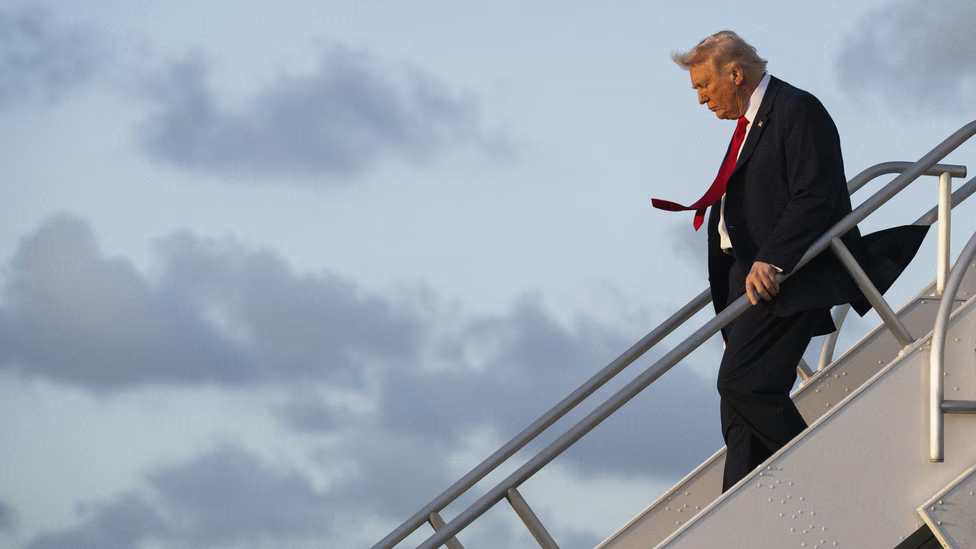 Donald Trump descending airplane stairs with red tie flying in the wind.