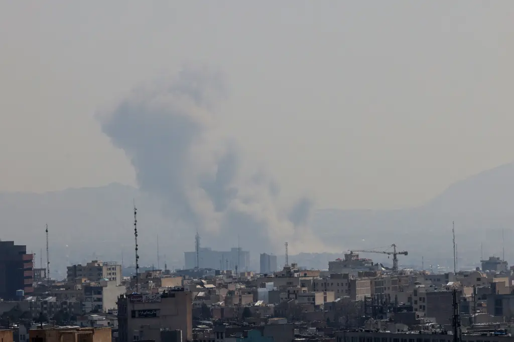 A daytime cityscape view under a hazy sky, showing a large plume of grey and white smoke rising from a building complex in the distance. Several telecommunication towers and a construction crane are visible among the city buildings.