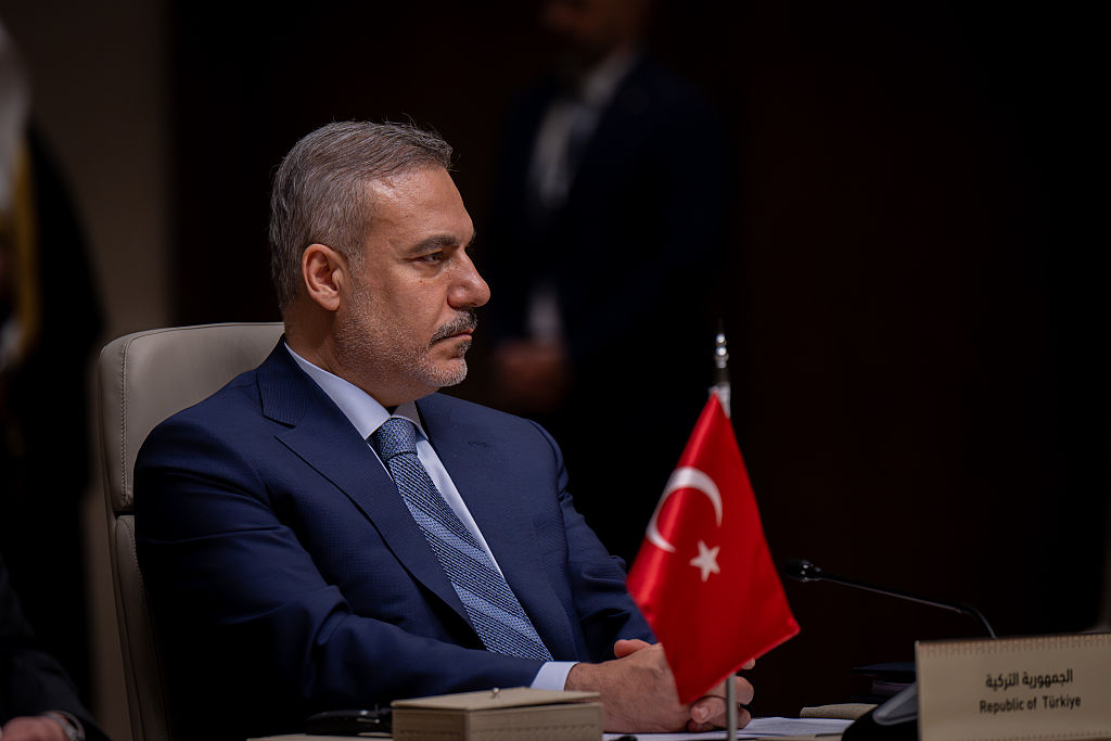 Hakan Fidan sitting at a diplomatic conference table with a Turkish flag in the foreground.