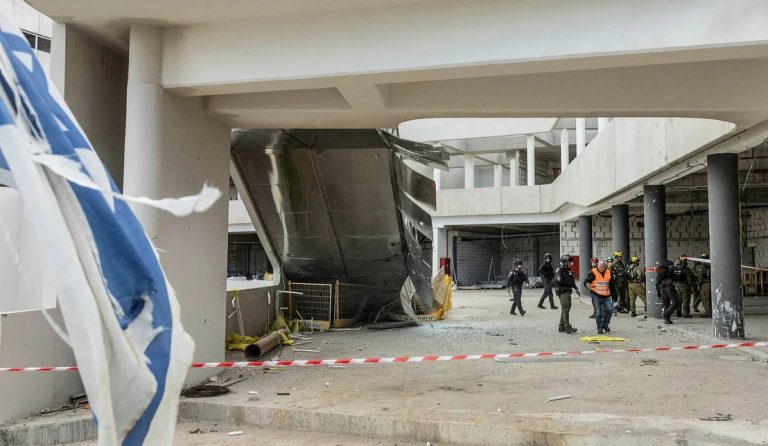 A tattered Israeli flag overlooks a damaged building interior where a large metal structure has collapsed, with emergency responders in orange vests and armed security personnel walking through the debris behind red and white caution tape.