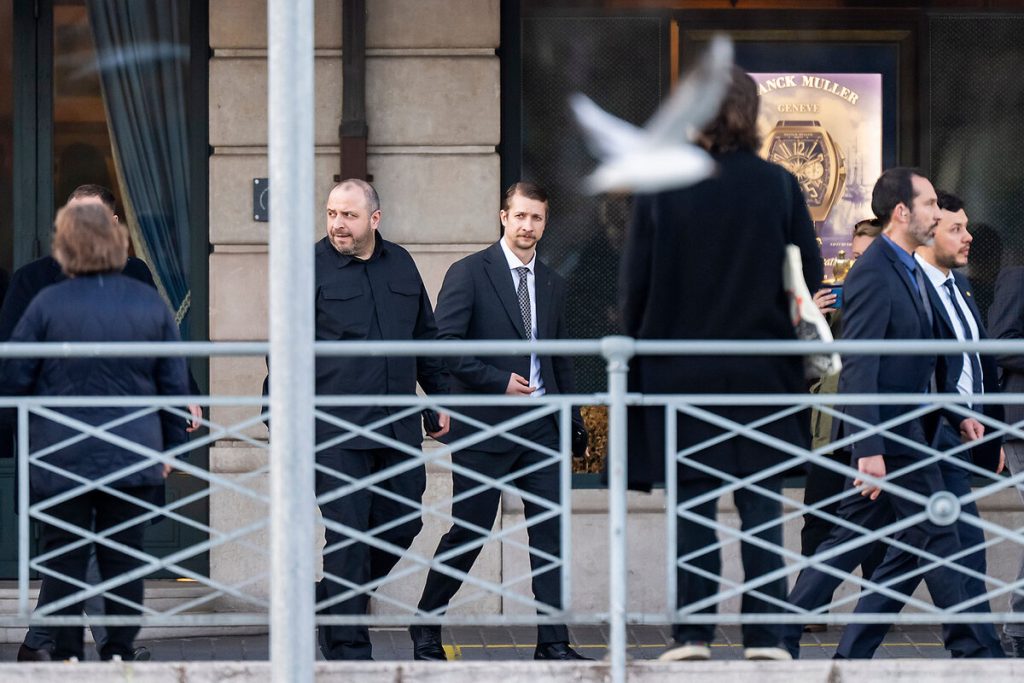 Several men in dark suits walking past a building with a "FRANCK MULLER" watch advertisement in the window, with a seagull blurred in flight.