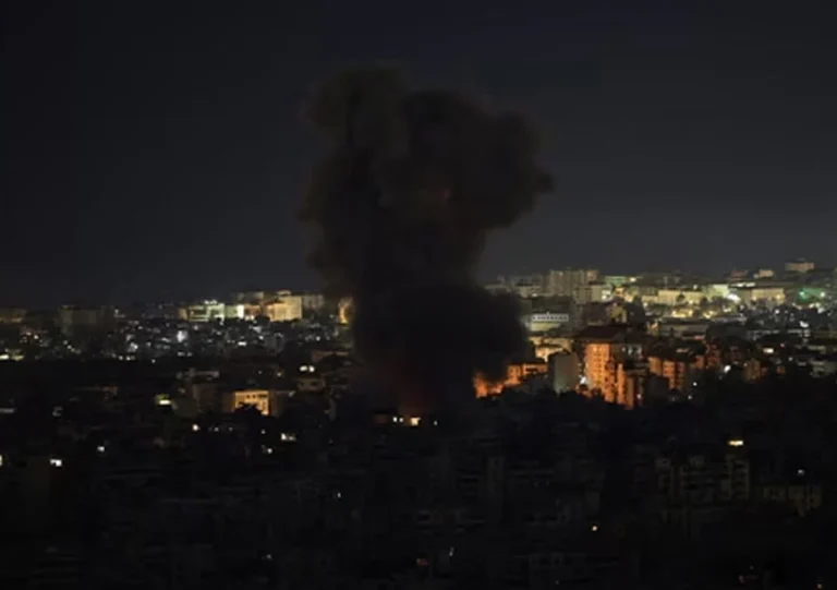 A nighttime view of a city skyline with a large, dark plume of smoke and fire rising from the center. The city lights are visible across the horizon under a dark sky.