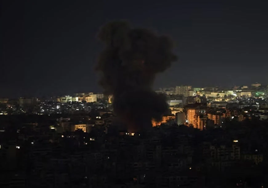 A nighttime view of a city skyline with a large, dark plume of smoke and fire rising from the center. The city lights are visible across the horizon under a dark sky.