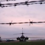 A military bomber aircraft on an airfield at dusk, viewed through a silhouette of barbed wire.