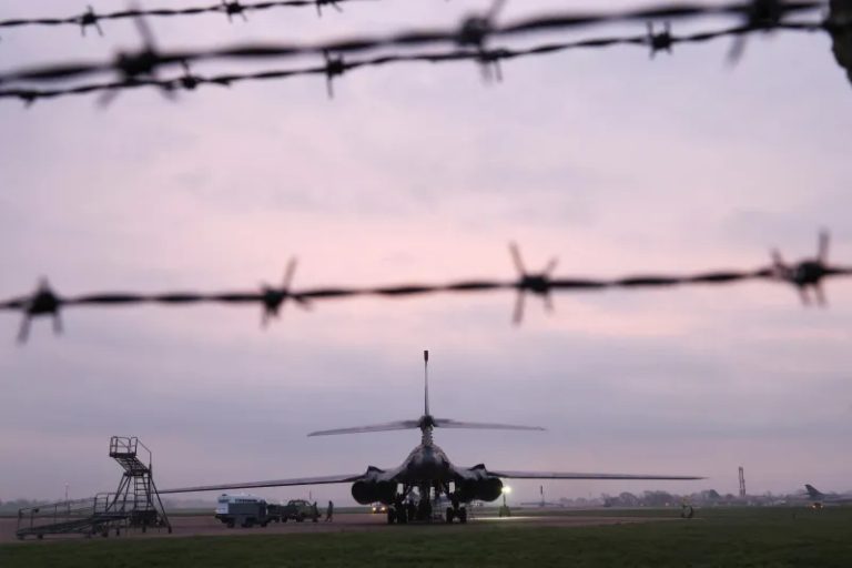 A military bomber aircraft on an airfield at dusk, viewed through a silhouette of barbed wire.