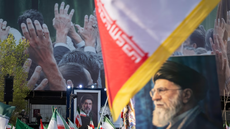 Large outdoor gathering in Iran featuring portraits of Ayatollah Khamenei and Ebrahim Raisi with national flags and a crowd background.