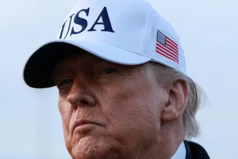 Close-up portrait of Donald Trump wearing a white "USA" hat with an American flag on the side.