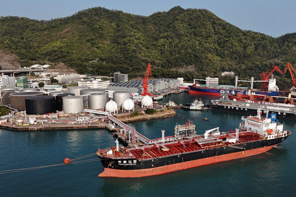 Large oil tankers and cargo ships docked at a major industrial port with storage tanks and green mountains in the background.