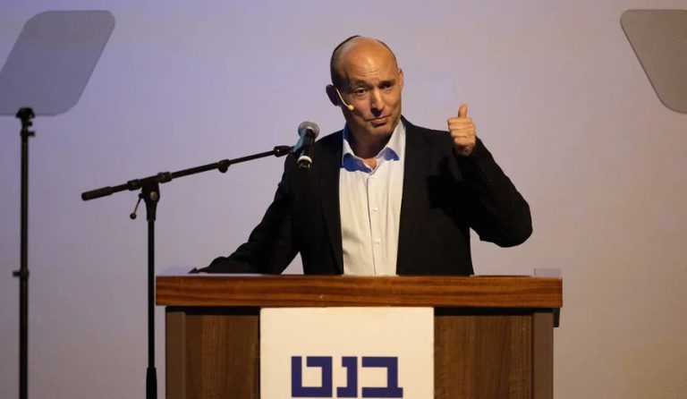 Naftali Bennett standing at a wooden podium with a microphone, giving a thumbs-up gesture during a political speech with a plain background.