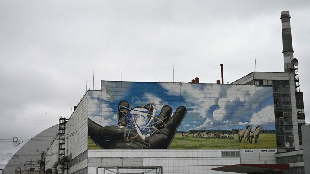 A large mural on the side of a Chernobyl Power Plant building depicting a hand holding an atom, with wild horses running in a green field.