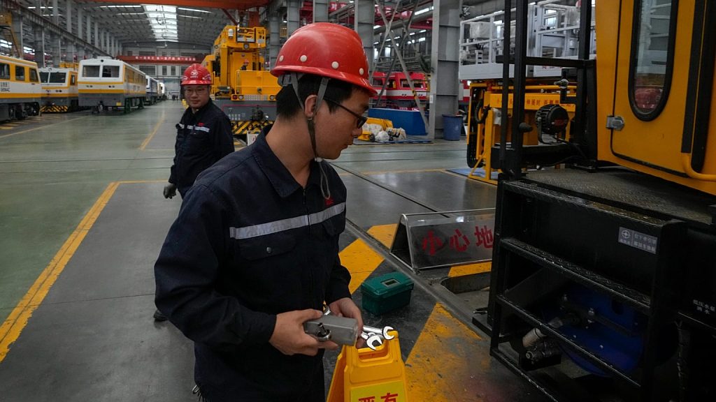 Chinese factory workers in red hard hats inspecting industrial machinery in a large manufacturing facility.
