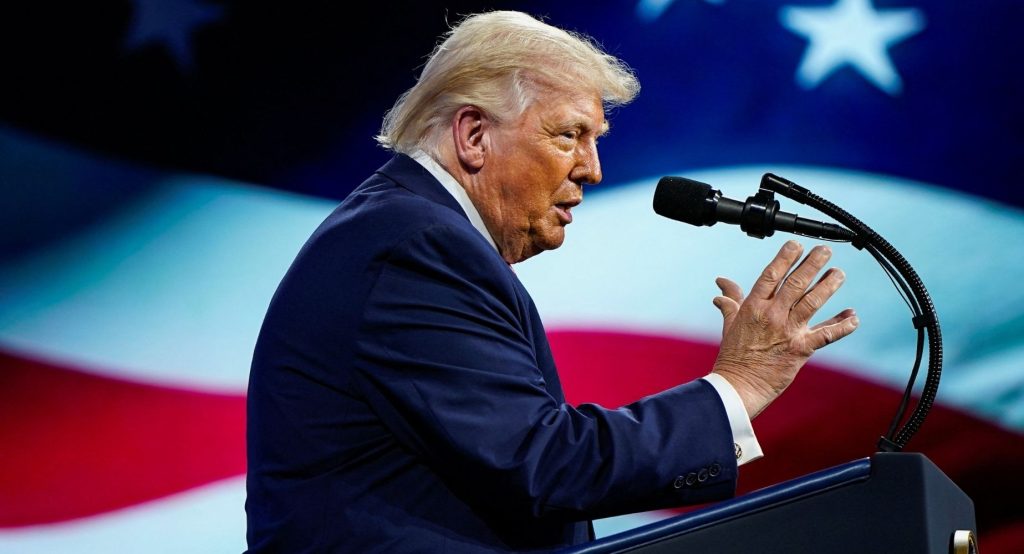 Donald Trump speaking at a podium with a large, blurred American flag in the background.