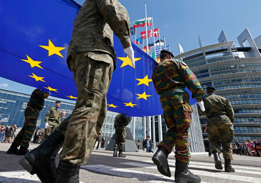 Soldiers in military uniforms carrying a large European Union flag in front of the European Parliament building with various national flags in the background.