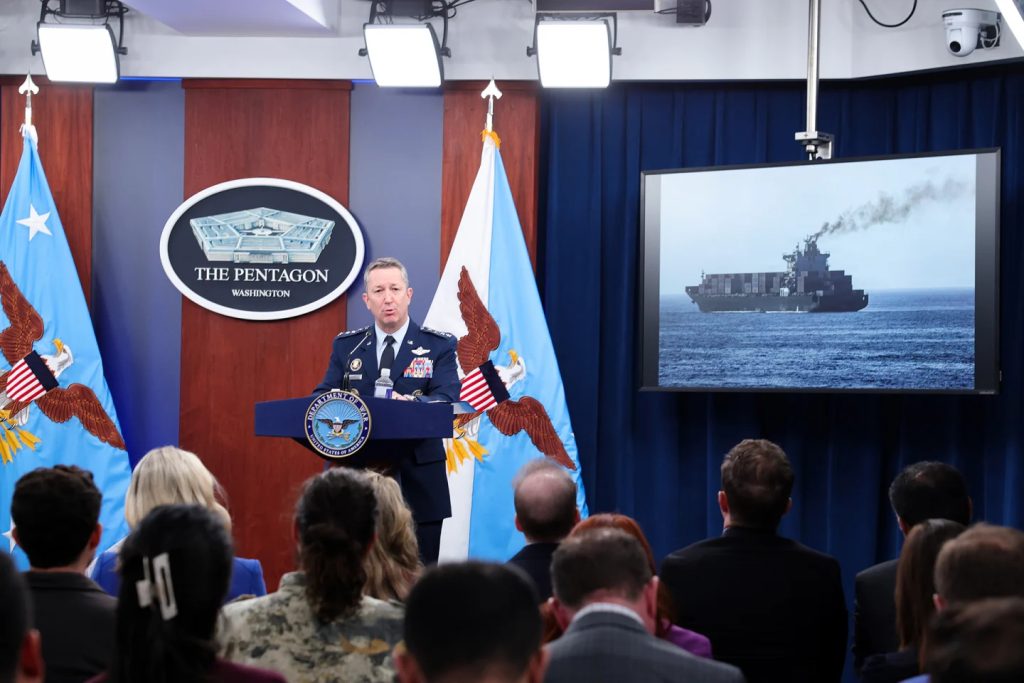 A U.S. military official speaking at a Pentagon press briefing with a screen showing a large container ship emitting smoke at sea.