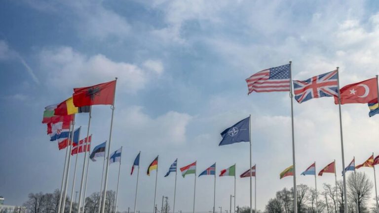 A circle of international flags, including the US, UK, and NATO, flying against a bright, cloudy sky.