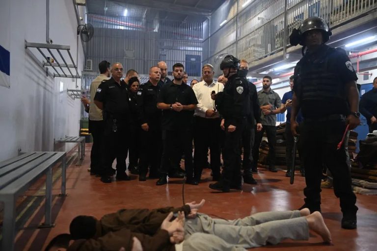 Itamar Ben-Gvir and Israeli security officials standing in a detention facility while several detainees lie face down on the floor with their hands restrained behind their backs.