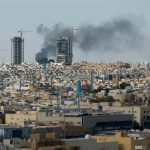 Panoramic view of a Middle Eastern city skyline with smoke rising from buildings under construction.