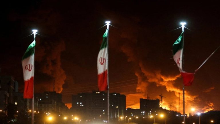 Iranian flags flying at night with a skyline filled with fire and thick smoke in the background.