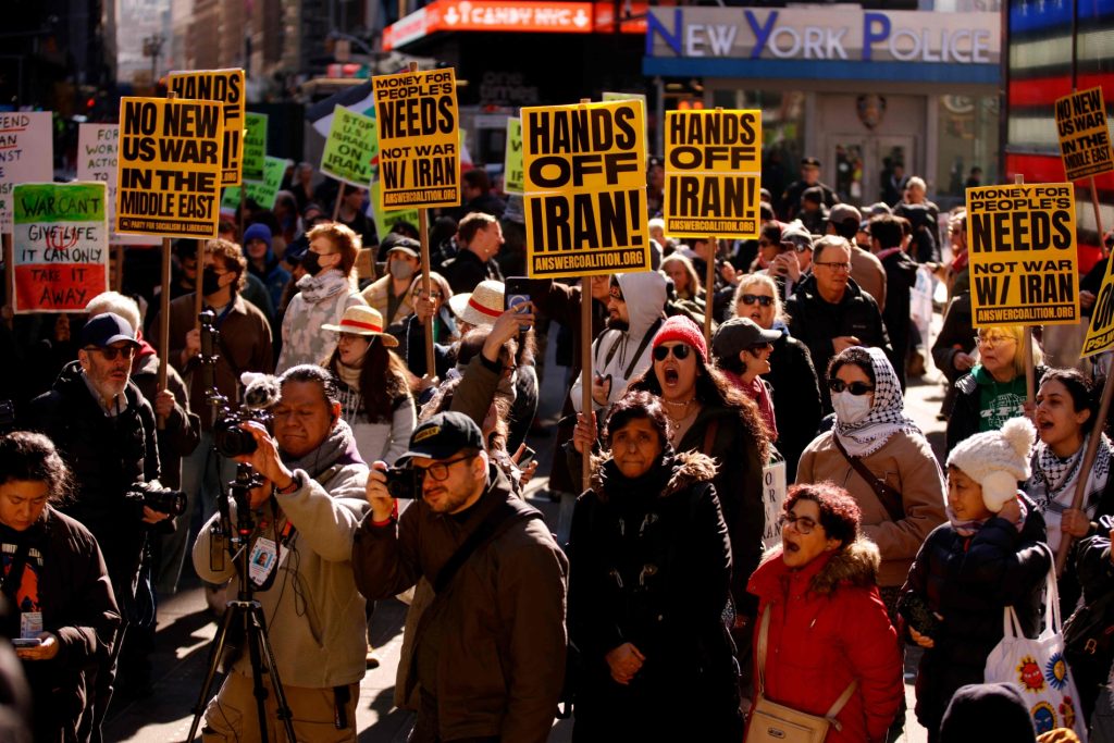 A crowded anti-war protest in Times Square, New York City, with demonstrators holding yellow signs that read "HANDS OFF IRAN!" and "NO NEW US WAR IN THE MIDDLE EAST!" while photographers capture the scene.