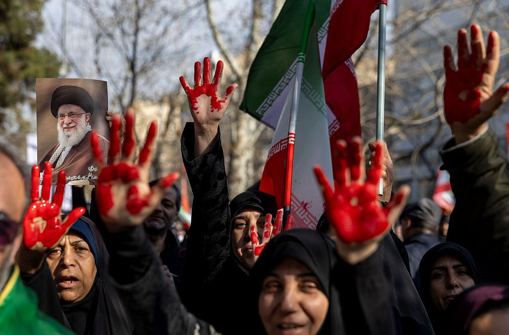 Iranian protesters in a crowd with red-painted palms raised and a portrait of Ali Khamenei.