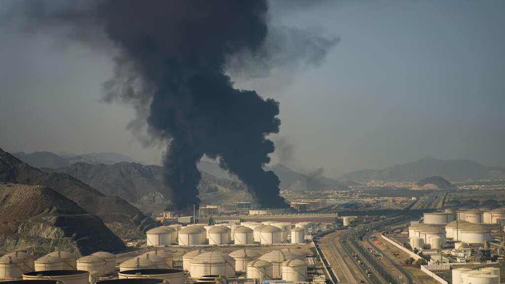 Thick black smoke rising from an industrial oil facility near mountains and a highway.