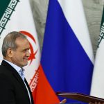 A close-up shot of a high-ranking official smiling while standing next to Vladimir Putin with Iranian and Russian flags in the background.