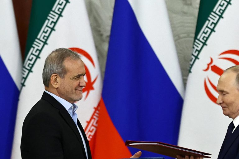 A close-up shot of a high-ranking official smiling while standing next to Vladimir Putin with Iranian and Russian flags in the background.