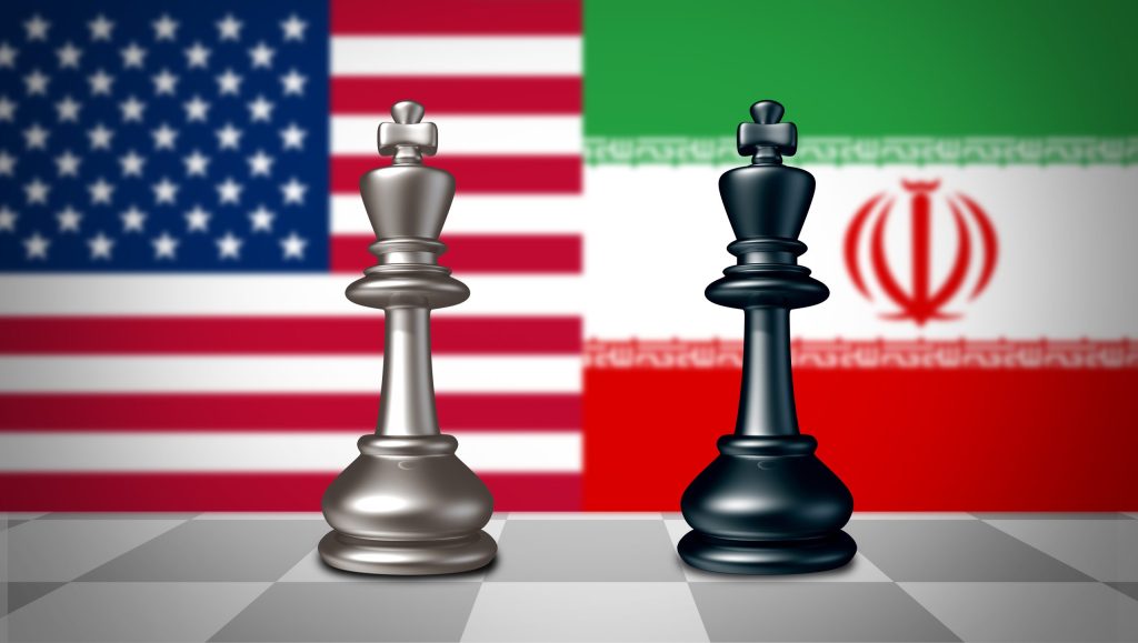 Two chess kings, one silver and one black, standing on a chessboard with the blurred flags of the United States and Iran in the background.