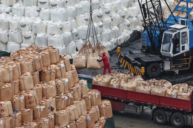 Industrial scene at a port showing a crane lifting large white and brown bags of sulfur.