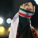 A raised clenched fist wrapped in the Iranian flag against a dark, blurred background with bokeh lights.