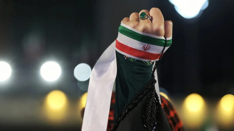 A raised clenched fist wrapped in the Iranian flag against a dark, blurred background with bokeh lights.