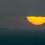 Silhouette of a large cargo ship on the ocean at sunset with a massive orange sun partially covered by clouds.