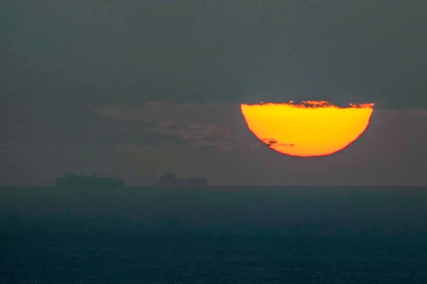 Silhouette of a large cargo ship on the ocean at sunset with a massive orange sun partially covered by clouds.