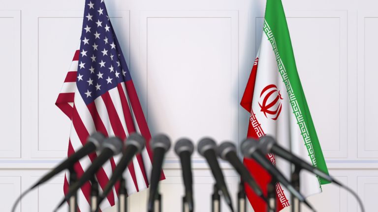 USA and Iran flags standing side-by-side behind a row of press microphones.