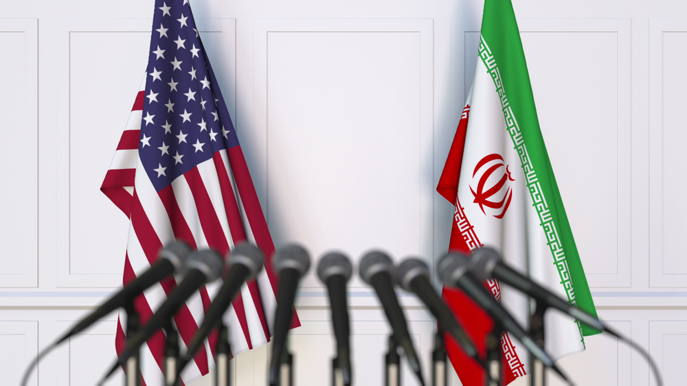 USA and Iran flags standing side-by-side behind a row of press microphones.