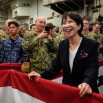 A high-ranking Japanese official smiling and engaging with military personnel on a naval vessel decorated with flags.