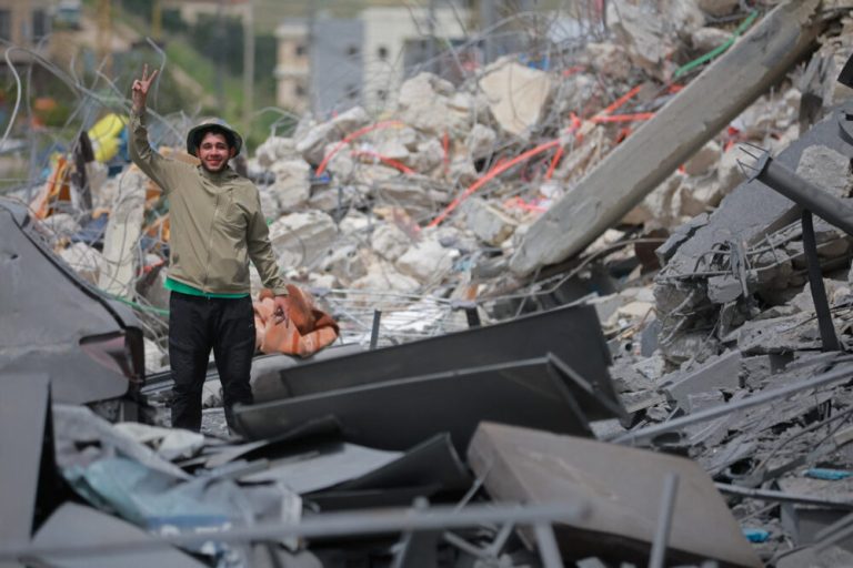 A young man standing amidst the grey rubble of destroyed buildings, flashing a peace sign with his hand.