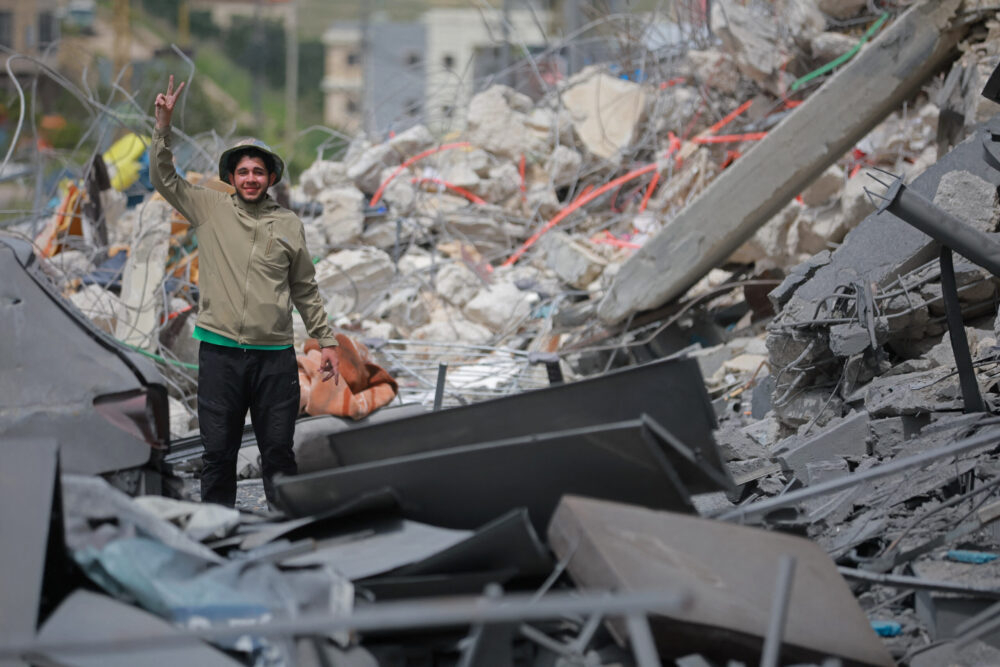 A young man standing amidst the grey rubble of destroyed buildings, flashing a peace sign with his hand.