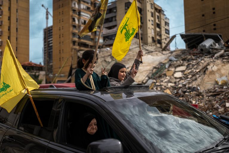 Children waving yellow Hezbollah flags from a car sunroof in front of destroyed buildings.