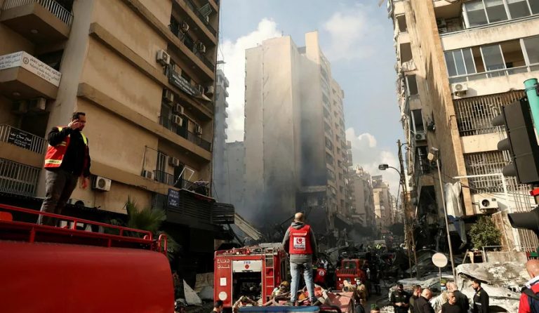 A street in Lebanon showing the aftermath of a military strike with smoke rising from damaged buildings and emergency responders on site.