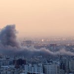 Thick grey smoke rising from a city skyline following an explosion or airstrike at dusk.