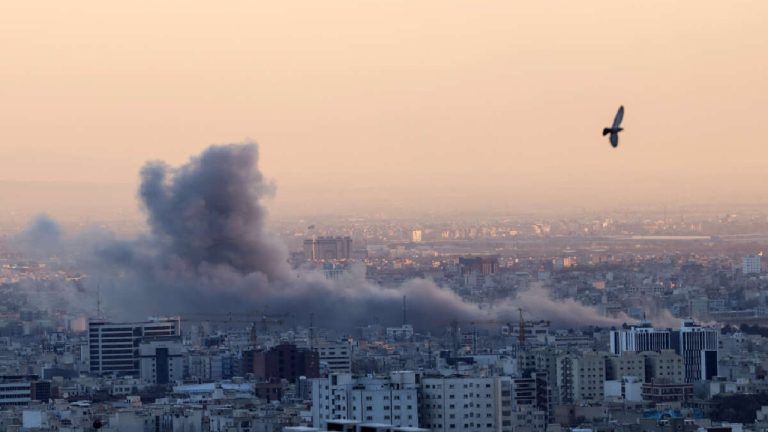 Thick grey smoke rising from a city skyline following an explosion or airstrike at dusk.