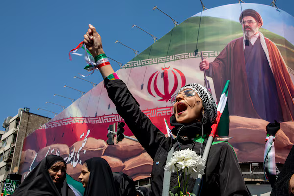 A woman in a kuffiyeh cheering in front of a large billboard featuring the Iranian flag and a portrait of a religious leader.
