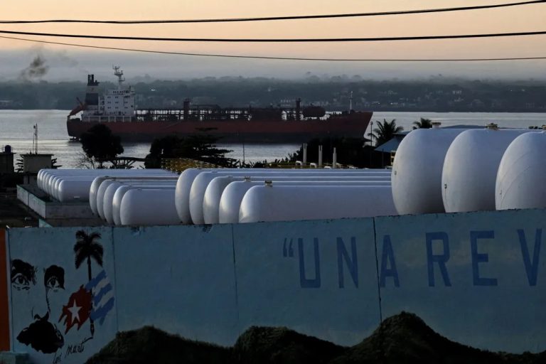 Large white fuel storage tanks in the foreground with a cargo ship docked in the distance at dusk.