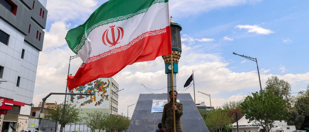 A large Iranian flag waving in an urban square with a man holding the flagpole.