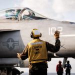 A deck crew member in a yellow safety vest signaling to a fighter jet pilot on an aircraft carrier.